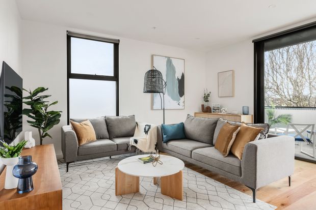 Bright contemporary living room with two grey sofas arranged around a round white coffee table, layered neutral and blue cushions, a geometric rug, timber flooring, indoor plants, framed abstract artwork, and large black-framed windows and glazing opening toward an outdoor balcony or terrace.