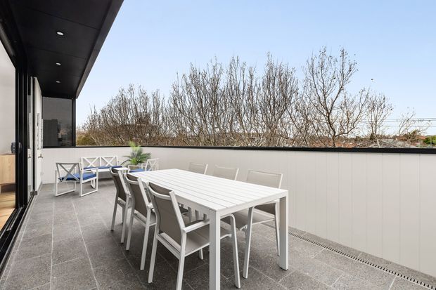 A contemporary rooftop or elevated outdoor dining terrace with a large white outdoor dining table and chairs, pale tiled flooring, a light privacy wall, and a dark overhanging soffit with recessed lighting. Glass doors connect the terrace to the interior, while leafless trees are visible beyond the parapet.