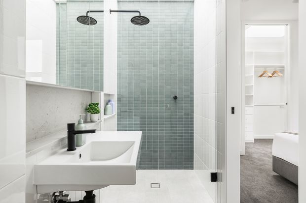 Bright contemporary bathroom with a wall-mounted white vanity and basin, black tapware, a large frameless shower enclosure, pale green-grey mosaic shower tiles, and white wall and floor tiles. A walk-in wardrobe and part of a bedroom are visible through the open doorway.