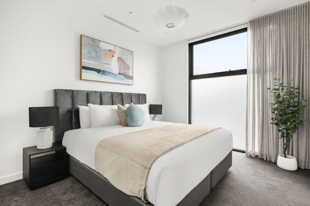 A bright, contemporary bedroom with a large upholstered bed, white bedding, a beige throw, dark bedside tables and lamps, a tall black-framed window, full-length curtains, carpeted flooring, abstract wall art, and a potted plant. The room has a clean, refined, neutral palette with soft natural light.
