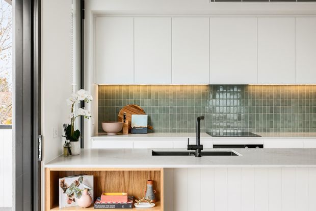 Bright contemporary kitchen with white flat-panel cabinetry, a pale stone island benchtop, and a glossy green-grey square-tile splashback illuminated by warm under-cabinet lighting. A black mixer tap and undermount sink sit at the island, while an open timber display niche adds warmth. Large glazing and a slim dark window frame bring natural light into the space.