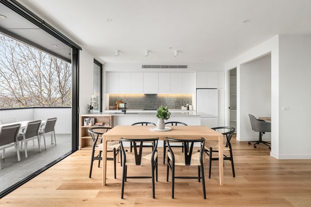 Bright open-plan dining and kitchen interior with a light timber dining table, black metal-framed chairs, white flat-panel cabinetry, a tiled backsplash, timber flooring, and large sliding glass doors opening to an outdoor area. The space has a clean, contemporary minimalist character with abundant natural light.