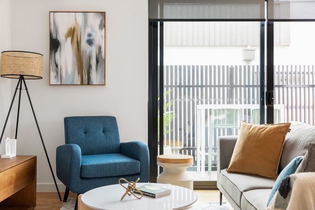 A bright, contemporary living room with a blue upholstered armchair, light sofa, round white coffee table, timber sideboard, tripod floor lamp, abstract artwork, and full-height black-framed glazing opening toward an outdoor balcony or terrace. The space has a restrained neutral palette with blue and ochre accents.