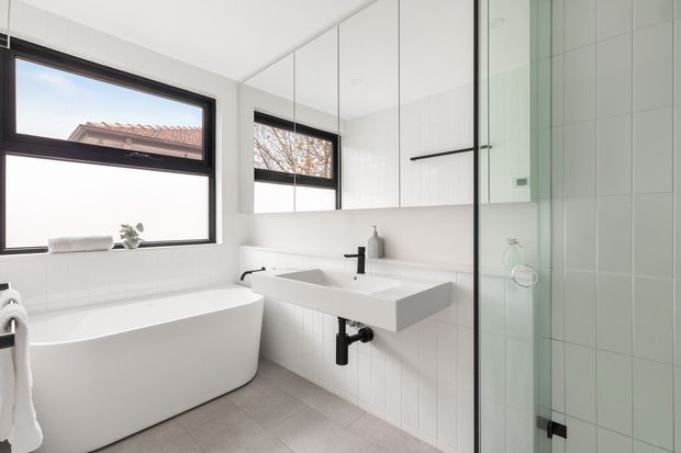 Bright contemporary bathroom with a freestanding white bathtub, wall-mounted white vanity basin, black tapware and hardware, large mirrored wall cabinets, white tiled walls, grey floor tiles, and a frameless glass shower enclosure. Natural light enters through dark-framed windows.