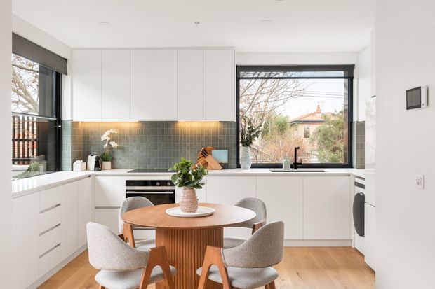 Bright contemporary open-plan kitchen and dining area with white flat-panel cabinetry, a muted green tiled splashback, timber flooring, and a round timber dining table surrounded by upholstered chairs. Large black-framed windows bring in natural light and overlook suburban homes and trees. Integrated appliances and under-cabinet lighting create a clean, refined fit-out.