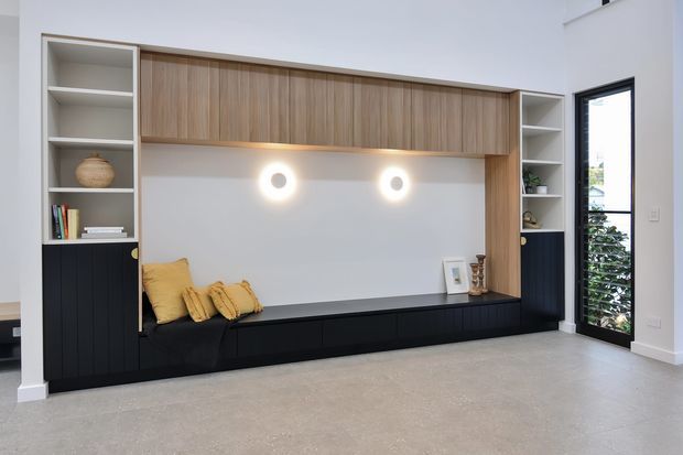 A bright, contemporary built-in living room feature wall with a long black low cabinet and bench seat, integrated open shelving, warm timber overhead cabinetry, and a white recessed backdrop. Two circular wall lights illuminate the seating nook, which is styled with mustard cushions and a few books and decorative objects. A tall glazed door or window is visible at right, and the room has a polished concrete or stone-look floor.