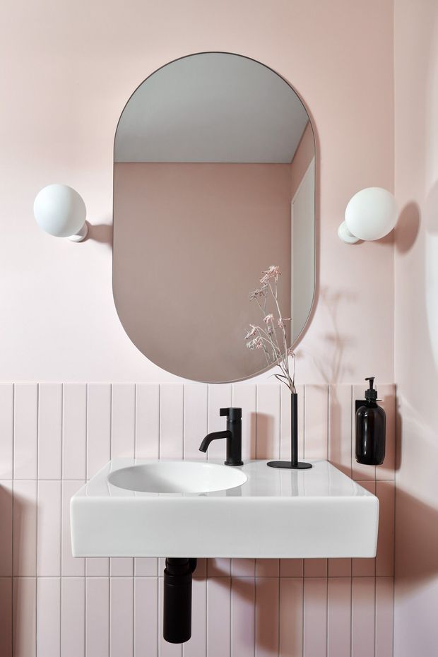 A compact, carefully styled bathroom vanity with a wall-mounted white basin and integrated pale countertop, set against blush-pink vertically arranged wall tiles. An oversized rounded-arch mirror dominates the wall, flanked by spherical white wall sconces. Matte black tapware, exposed plumbing, soap dispenser, and a minimalist vase with dried stems provide dark and organic accents. The composition is bright, refined, and contemporary with a soft pastel palette.