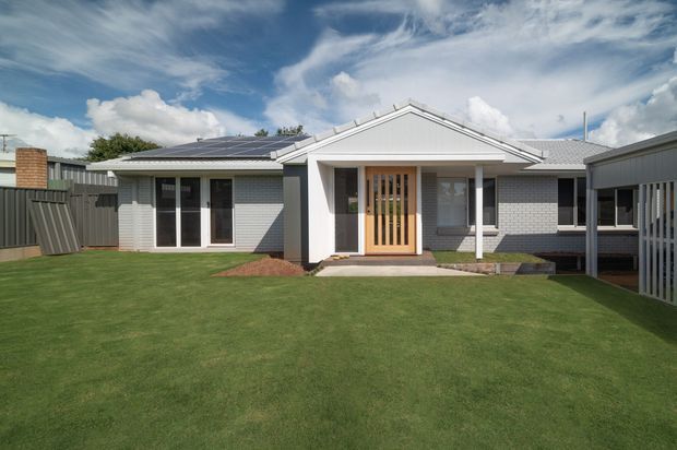 Single-storey suburban home viewed from the front across a neatly maintained lawn. The house has a light rendered or painted brick facade, a gabled roof, a central entry with a timber slatted door, large glazed openings, a solar-panelled roof section, and a covered carport or side structure on the right. The palette is predominantly light grey, white, charcoal, and natural timber.
