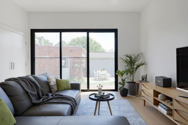 Bright contemporary living room with a large black-framed glazed door opening to a small outdoor courtyard. The room features a black leather sectional sofa with layered throws and cushions, a round coffee table, pale area rug, indoor plants, a light timber media console, and a wall-mounted television. White walls and ceiling create an airy, uncluttered interior.