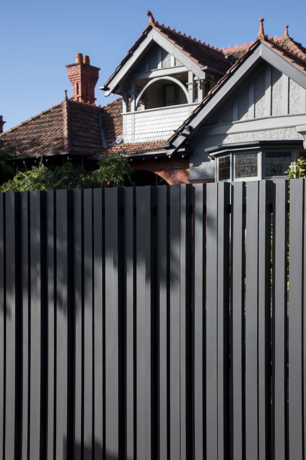 Exterior view of a heritage-style residential house behind a tall dark vertical-slat fence. The house features steep gabled roofs, terracotta tiled roofing, decorative timber gable detailing, a projecting balcony with white patterned cladding, ornate window trim, and a brick chimney. Clear blue sky and garden planting are visible around the building.
