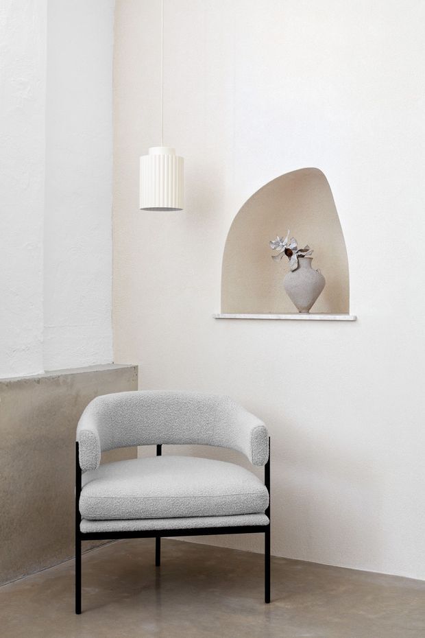 A serene minimalist interior corner featuring a textured white plaster wall, a sculptural arched recessed niche with a ceramic vase, a hanging ribbed pendant light, and a contemporary upholstered armchair with a black metal frame. The floor and low half-height wall are finished in smooth warm grey concrete or microcement.