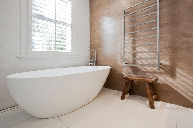 Bright contemporary bathroom featuring a freestanding white oval bathtub, white tiled flooring and wall, a warm wood-look feature wall with a chrome heated towel rail, and a small rustic timber stool. Plantation shutters provide filtered natural light.