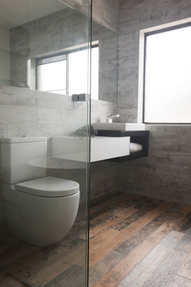 A modern bathroom viewed through a frameless glass shower screen, featuring a wall-hung white toilet, floating black vanity with a white rectangular basin, large-format grey textured wall tiles, a full-height mirror, and weathered timber-look flooring. A high window provides bright natural light.