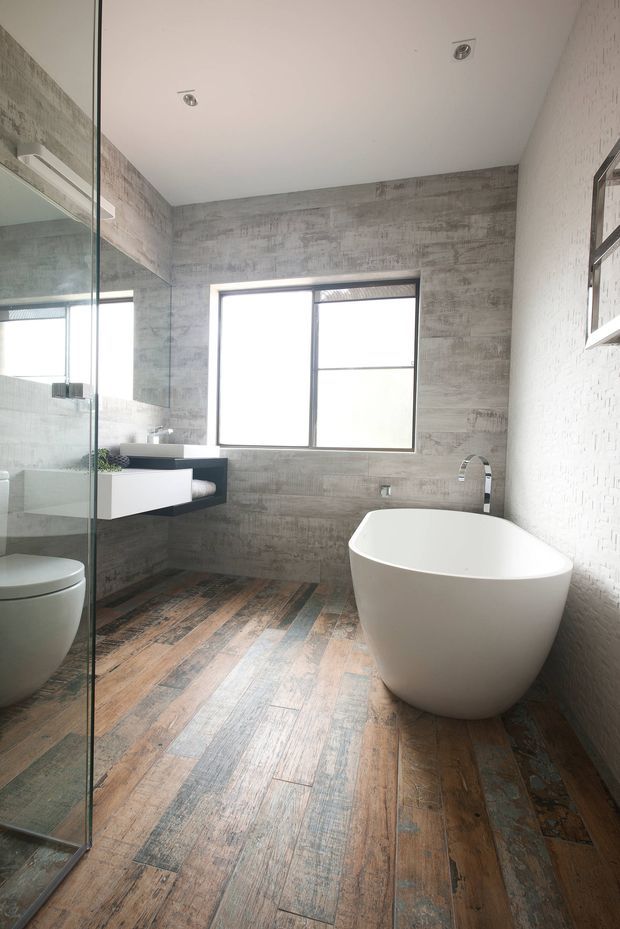 Contemporary bathroom with a freestanding white bathtub, wall-mounted vanity, glass-enclosed shower, and toilet. The room features grey textured wall tiles, a weathered timber-look floor, black-framed glazing, recessed ceiling lights, and a large mirror creating a bright, minimalist atmosphere.