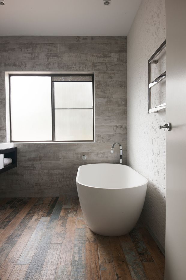 A contemporary bathroom viewed from the doorway, featuring a freestanding white bathtub, wall-mounted chrome tapware, a recessed mirrored cabinet, a frosted black-framed window, textured white wall tiles, and weathered grey-brown timber-look flooring. The room has a restrained industrial feel with exposed concrete-effect wall tiling and mixed natural materials.