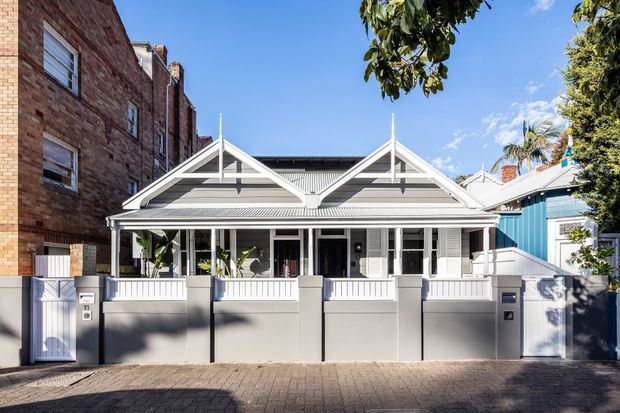 Street-facing view of a renovated single-storey terrace or bungalow-style residence with a symmetrical gabled roof, decorative timber gable detailing, broad front veranda, pale rendered boundary wall, and light weatherboard cladding. A taller brick building adjoins the left side, while mature trees and a neighbouring blue-clad house frame the property.