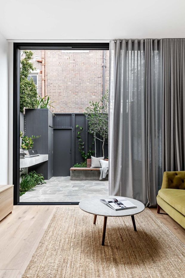 A bright, contemporary living room opening onto a compact landscaped courtyard through a large black-framed glazed door. The room features pale timber flooring, a large natural woven rug, floor-to-ceiling grey curtains, a round light stone coffee table, and a mustard-green upholstered armchair. The courtyard includes grey paving, dark vertical cladding, built-in bench seating, and established planting, with a brick wall beyond.