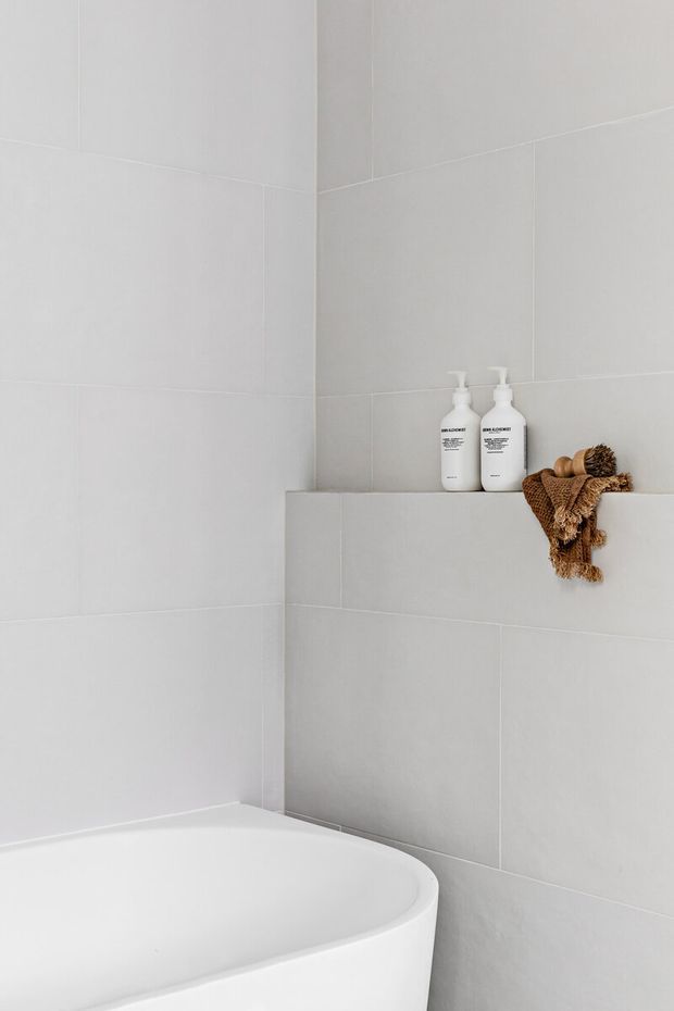 Close-up view of a minimalist bathroom corner featuring a white freestanding bathtub, large light-grey wall tiles, and a recessed tiled shelf holding white toiletries and a folded brown textured towel.