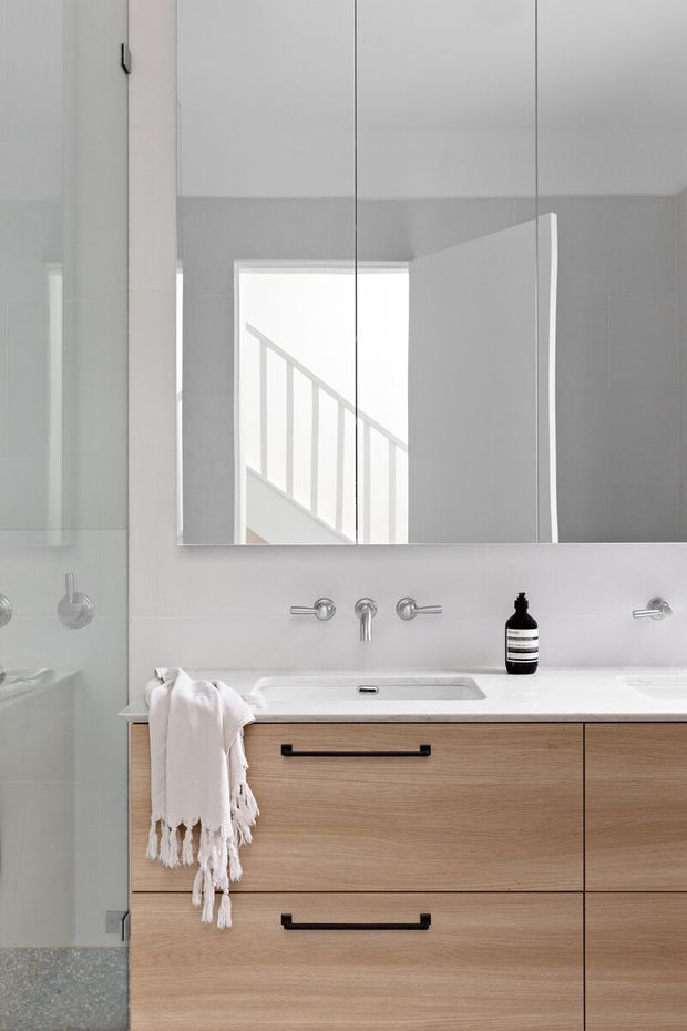 Minimal, light-filled bathroom vanity with a large frameless mirrored cabinet, pale tiled walls, integrated white basin, wall-mounted chrome tapware, and warm light-wood drawer cabinetry with black pulls. A glass shower enclosure is visible at left, and a stairwell is reflected in the mirror.