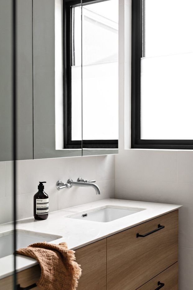 Contemporary bathroom vanity with a white integrated basin and benchtop, wall-mounted chrome tapware, warm timber drawers with black pulls, pale tiled walls, and black-framed windows. A large mirror reflects the vanity and window, while a dark grey wall panel and neutral textiles add contrast.