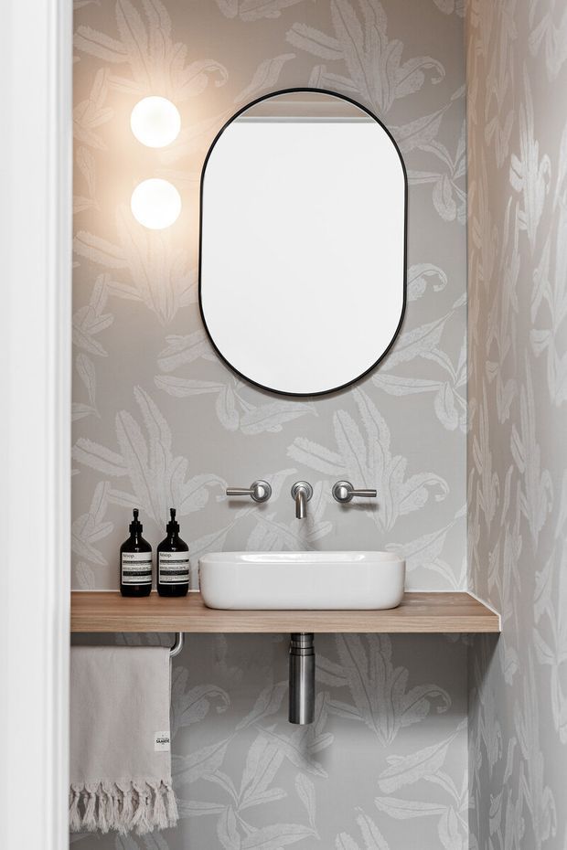 A compact powder room with a wall-mounted white vessel sink on a warm timber shelf, exposed chrome plumbing, and a brushed metal wall-mounted tap set. Grey wallpaper with a large white botanical pattern covers the walls, complemented by a rounded black-framed mirror and two globe wall lights. A hand towel and dark soap bottles sit on the shelf.
