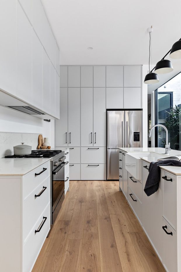 Bright contemporary galley kitchen with extensive white flat-panel cabinetry, black handles, stainless-steel appliances, white benchtops, timber-look flooring, and black-and-white pendant lighting. A window and glazed door bring in natural light and show greenery outside.