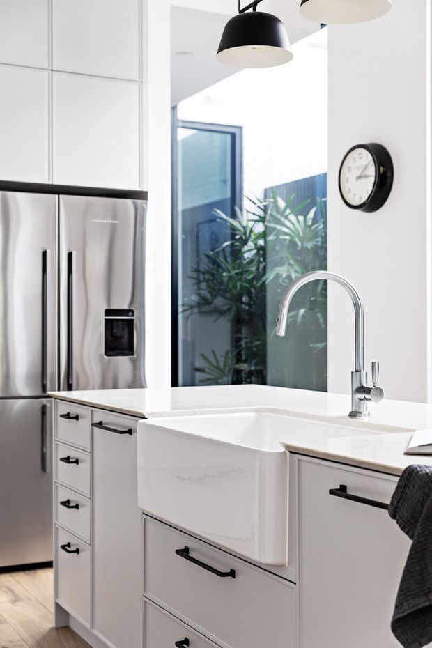 Bright contemporary kitchen featuring white shaker-style cabinetry, a large stainless-steel French-door refrigerator, a deep white farmhouse apron-front sink, pale benchtops, black pendant lights, and matte black hardware. A tall window provides views of planted greenery and an adjacent wall, while a round wall clock adds a functional accent.