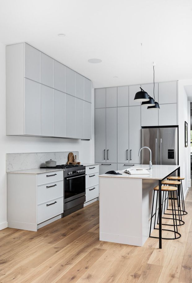 Bright contemporary kitchen with white floor-to-ceiling cabinetry, a marble-look splashback and island benchtop, stainless-steel appliances, timber flooring, and a black pendant-light and stool arrangement. The compact open-plan layout has a clean, refined appearance with ample built-in storage.