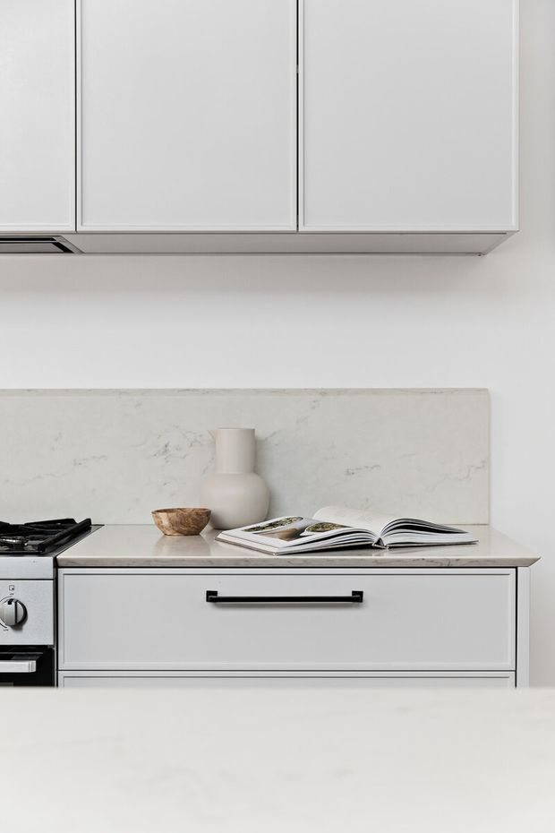 A close-up view of a bright, minimalist kitchen with white Shaker-style cabinetry, black linear handles, a pale stone benchtop and matching marble-look splashback. A freestanding black-and-stainless steel cooker is partially visible at left, while an open cookbook and neutral ceramic and wooden vessels provide subtle styling.