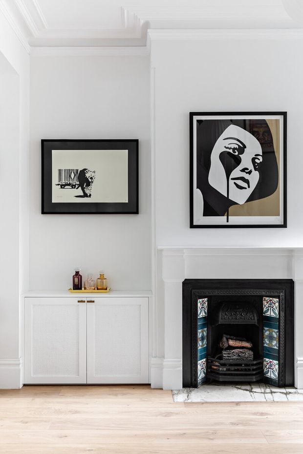 A refined white traditional interior featuring ornate ceiling mouldings, a decorative white mantel and a restored black cast-iron fireplace with colourful tiled detailing. A built-in white cabinet sits beside the fireplace, topped with small decorative objects, while two monochrome framed artworks hang above. Pale timber flooring and abundant white finishes create a bright, elegant room.