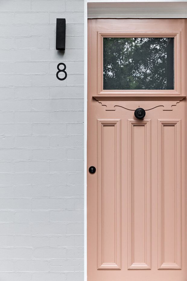 Close-up exterior view of a traditional panelled front door painted dusty blush pink, set beside a white-painted brick wall. The door has ornate raised mouldings, a small upper glazed panel, dark circular hardware, a black wall light and the house number 8.