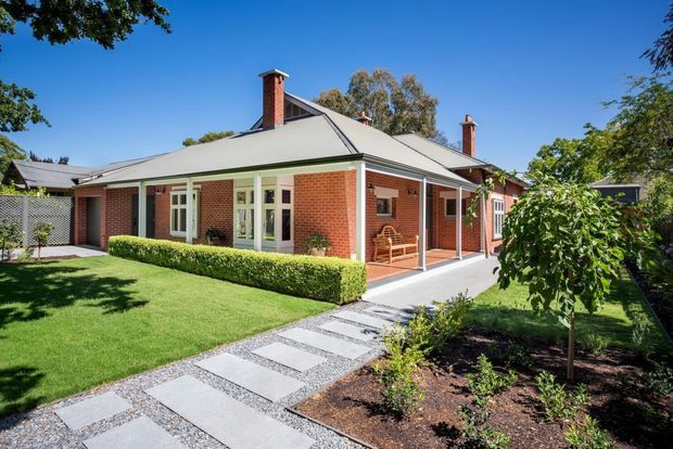 A single-storey traditional brick home with a broad hipped metal roof, multiple brick chimneys, cream-painted timber-framed windows and doors, and a covered verandah. The house is surrounded by a neatly landscaped front garden with clipped hedges, lawn, garden beds, and a concrete stepping-stone path through gravel.