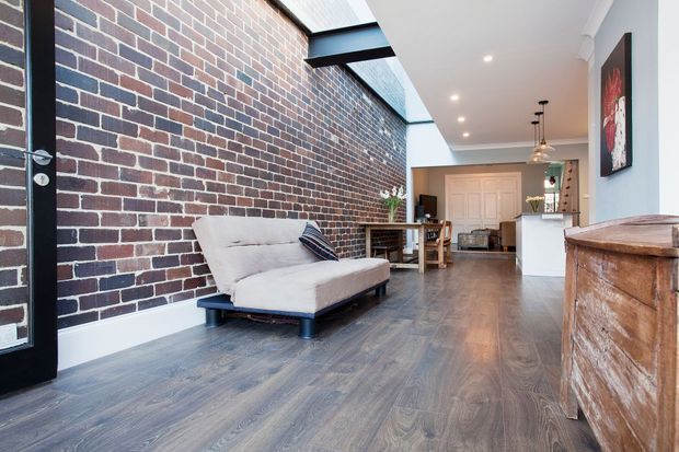 Open-plan industrial-style interior combining a living area, dining area and kitchen. A dark exposed brick feature wall runs along the left side beside a light sofa, while timber-look flooring leads toward a compact white kitchen with pendant lighting. The space has a bright, high ceiling with skylight-like glazing and a large rustic timber storage cabinet in the foreground.