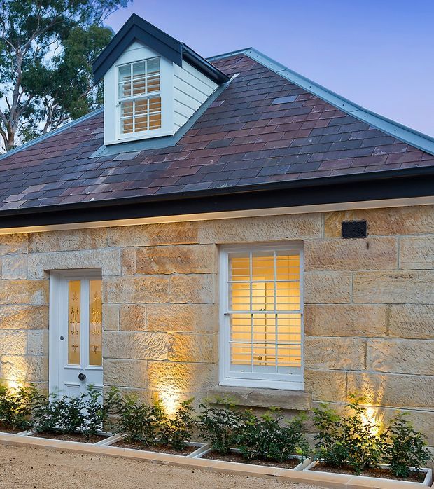 Exterior view of a heritage-style single-storey stone cottage with a steep pitched gable roof, dormer window, slate roof tiles, white-framed windows and glazed entry door. Warm wall lighting highlights the textured sandstone facade and landscaped planting beds along the front.