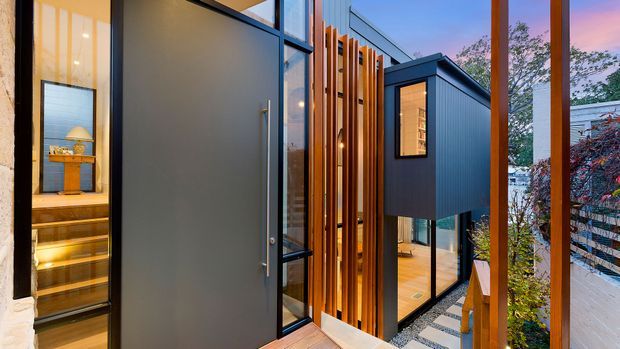 A contemporary multi-level residential exterior or entry approach with dark metal cladding, extensive glazing, warm timber vertical screens, and a timber deck. The view looks toward a glazed entrance and a compact projecting upper volume, with warm interior lighting visible through the windows and a landscaped boundary at the side.