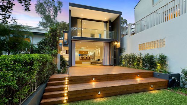 Two-storey contemporary rear extension viewed from a landscaped garden, featuring a dark-framed glazed facade, open-plan living area, upper balcony, and a broad timber deck with integrated step lighting.