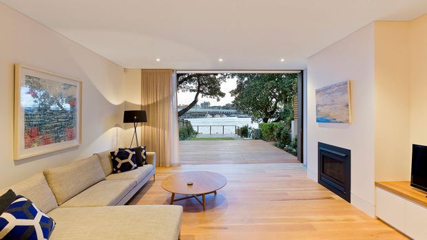 Contemporary living room with a large pale sectional sofa, round timber coffee table, recessed ceiling lights, artwork, and a built-in fireplace. Full-height glazing opens onto a timber deck and landscaped waterfront outlook, creating a bright indoor–outdoor connection.