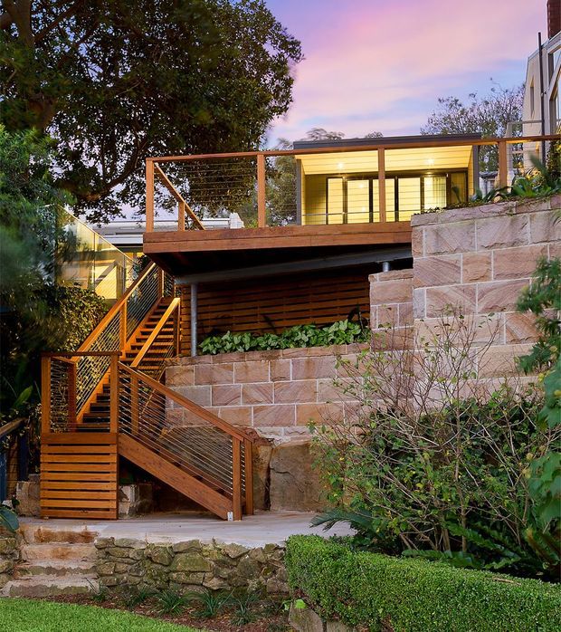 Exterior view of a multi-level contemporary residential addition set into a landscaped, sloping site. The image features warm timber decking and stairs with cable balustrades, a glazed upper-level room, horizontal timber screening, and substantial exposed sandstone retaining walls surrounded by mature planting.
