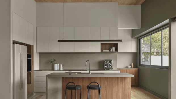 Contemporary kitchen interior with a central timber-front island and two bar stools, white flat-panel cabinetry, integrated stainless-steel refrigerator, neutral stone-look benchtops and splashback, and a long black linear pendant. A large window provides natural light, while the timber ceiling and olive-grey wall add warmth and contrast.