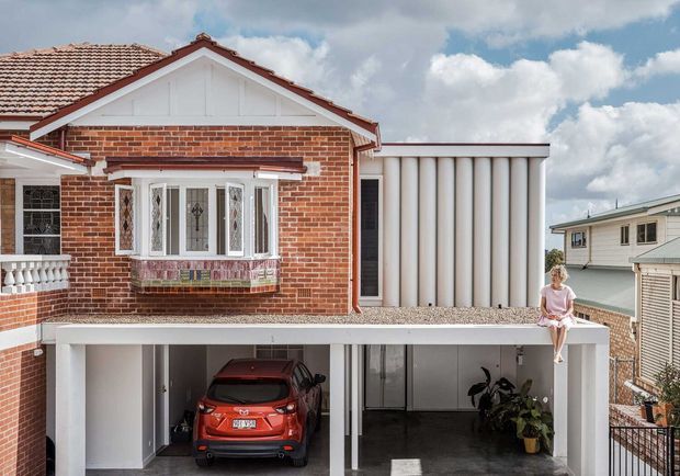 Exterior view of a compact two-storey brick dwelling with a preserved gabled street-facing facade, tiled pitched roof, decorative bay window, and a contemporary white addition. The lower level forms an open carport and covered entry area, with a red vehicle and potted plants visible beneath the structure. Neighbouring homes and a partly cloudy sky surround the property.