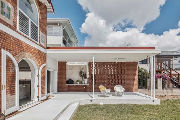 Exterior view of a brick residential home with a covered outdoor patio, arched openings, white louvred windows, and a flat-roofed verandah. The patio includes built-in brickwork, a small outdoor seating area, and a concrete terrace overlooking a lawn.