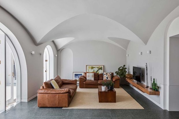 A bright, spacious living room with sculptural white arched and vaulted ceiling forms, tall arched windows and doors, brown leather sofas, a timber coffee table and media console, and a neutral area rug over dark flooring. The room has a refined contemporary character with strong curved architectural detailing.