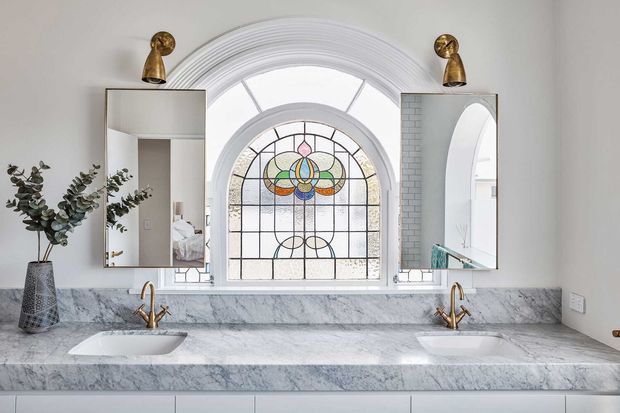 A refined double-sink bathroom vanity beneath a large arched stained-glass window. The wall and benchtop are clad in pale veined marble, with white undermount basins, brass tapware, arched mirrors, and warm brass wall lights. A bedroom is faintly visible through the left mirror reflection.