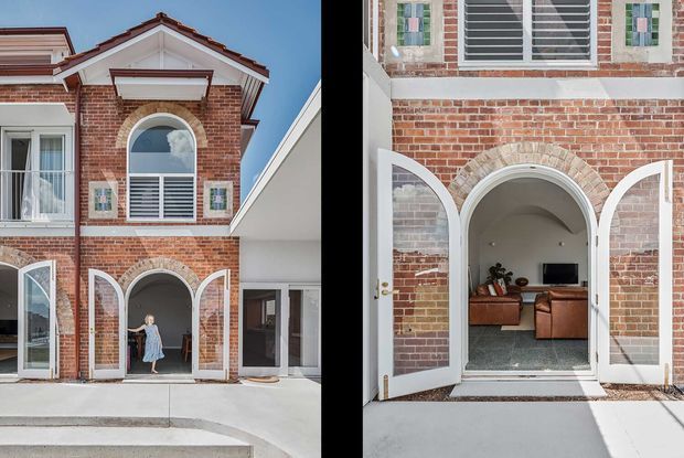 A red-brick multi-storey residential facade with white trim, arched openings, louvred windows, and a pitched tiled roof. The open arched doors reveal a living room beyond, with timber-toned seating and a television. The image presents both an exterior elevation and a closer view into the entry/living space.