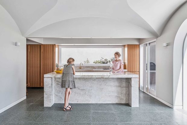 Bright contemporary kitchen with a large marble-clad island, integrated sink and tap, warm timber cabinetry, expansive glazing, and a sculptural vaulted white ceiling. Two people occupy the space, with a child standing at the island and an adult behind it.