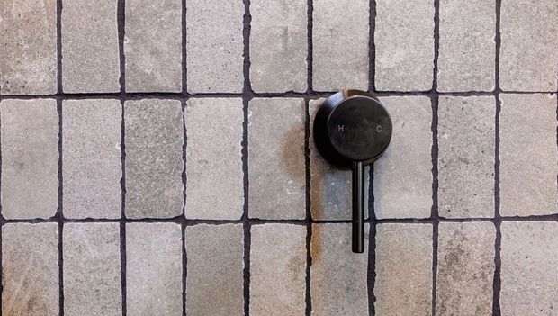 Close-up of a wall-mounted black circular control with a slim vertical handle, set against a grid of small textured light-grey tiles with dark grout lines. The image appears to show a shower or bathroom control fitting, with a muted, industrial material palette.