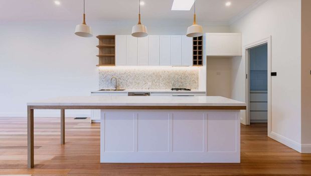 Bright contemporary kitchen featuring a large white island with panelled cabinetry and a timber-framed breakfast bar, white shaker-style wall cabinetry, open timber shelving, a patterned light backsplash, integrated appliances, and three pendant lights. Warm timber flooring and a neutral palette create a clean, inviting interior.