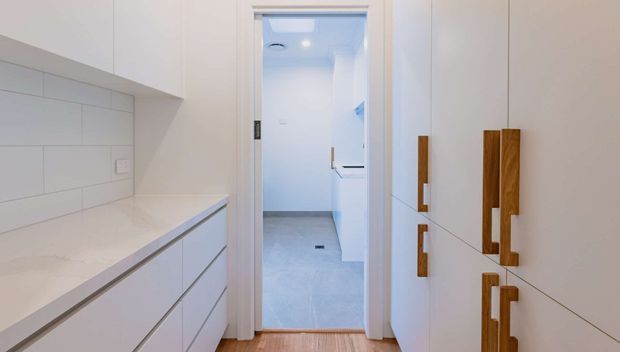 A bright, compact contemporary kitchen or butler’s pantry looking through an open doorway toward a tiled utility space. White flat-panel cabinetry and drawers line both sides, with long contrasting timber handles, a pale stone benchtop, and white rectangular wall tiling. The foreground has a warm timber floor, while the adjoining room has pale grey tile flooring.