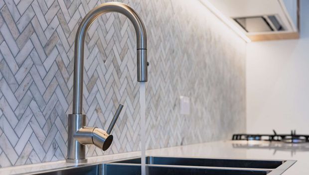 Close-up of a contemporary kitchen sink with a tall chrome gooseneck mixer tap and water running into a dark undermount sink. The backsplash features a full-height pale grey and white herringbone tile pattern, with a light benchtop, white cabinetry, and a gas cooktop visible in the background.
