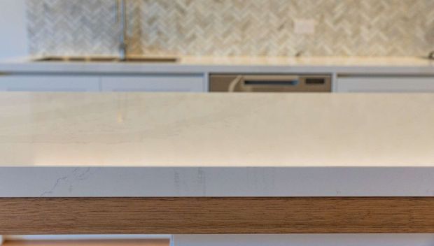 Close-up detail of a contemporary kitchen island, showing a pale stone benchtop with a thick waterfall-style edge, a warm timber panel below, pale cabinetry, and a textured chevron-patterned splashback in the background.